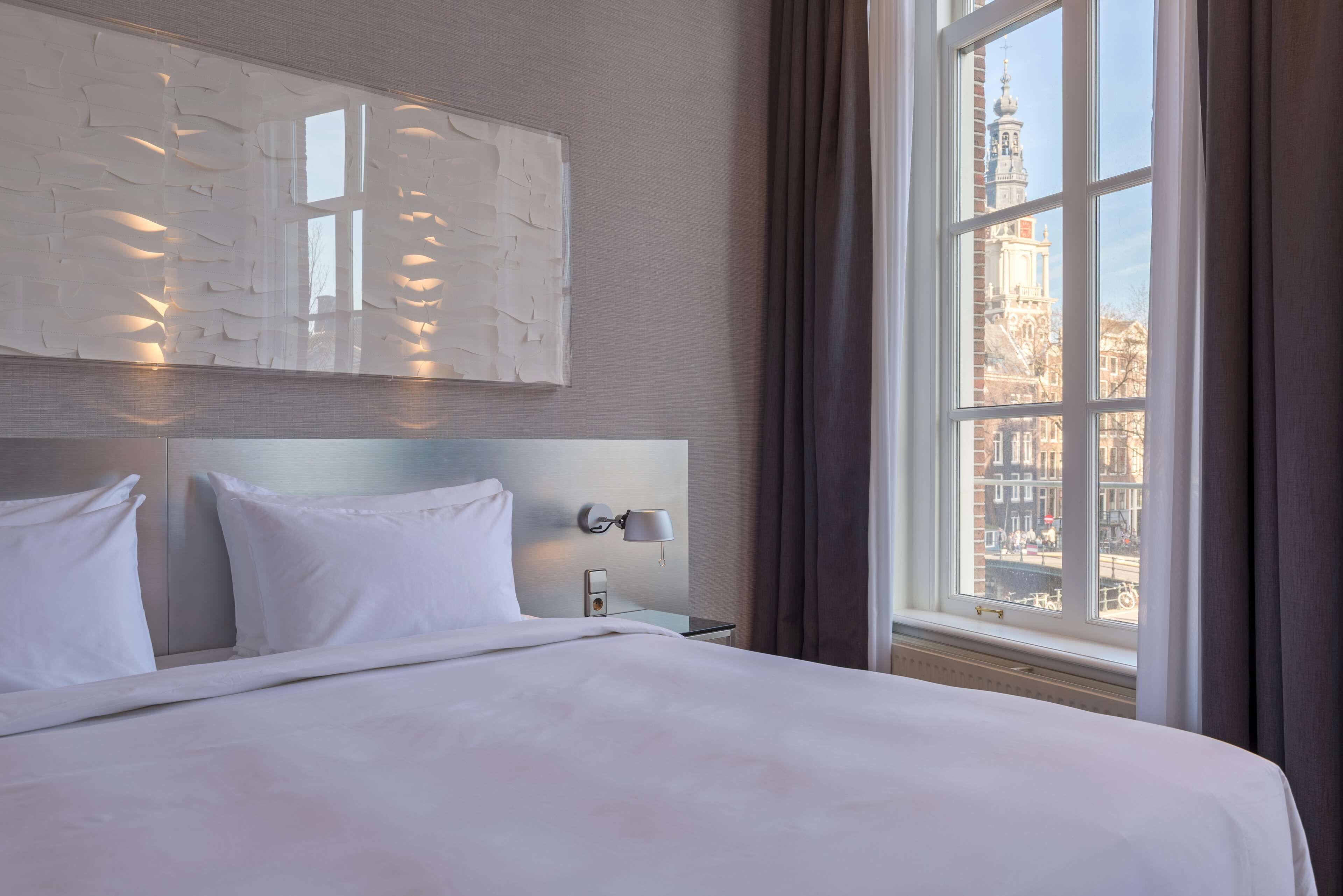 Hotelkamer met een groot bed, nachtkastje met lamp, wanddecoratie en een raam met uitzicht op een stadsbeeld.