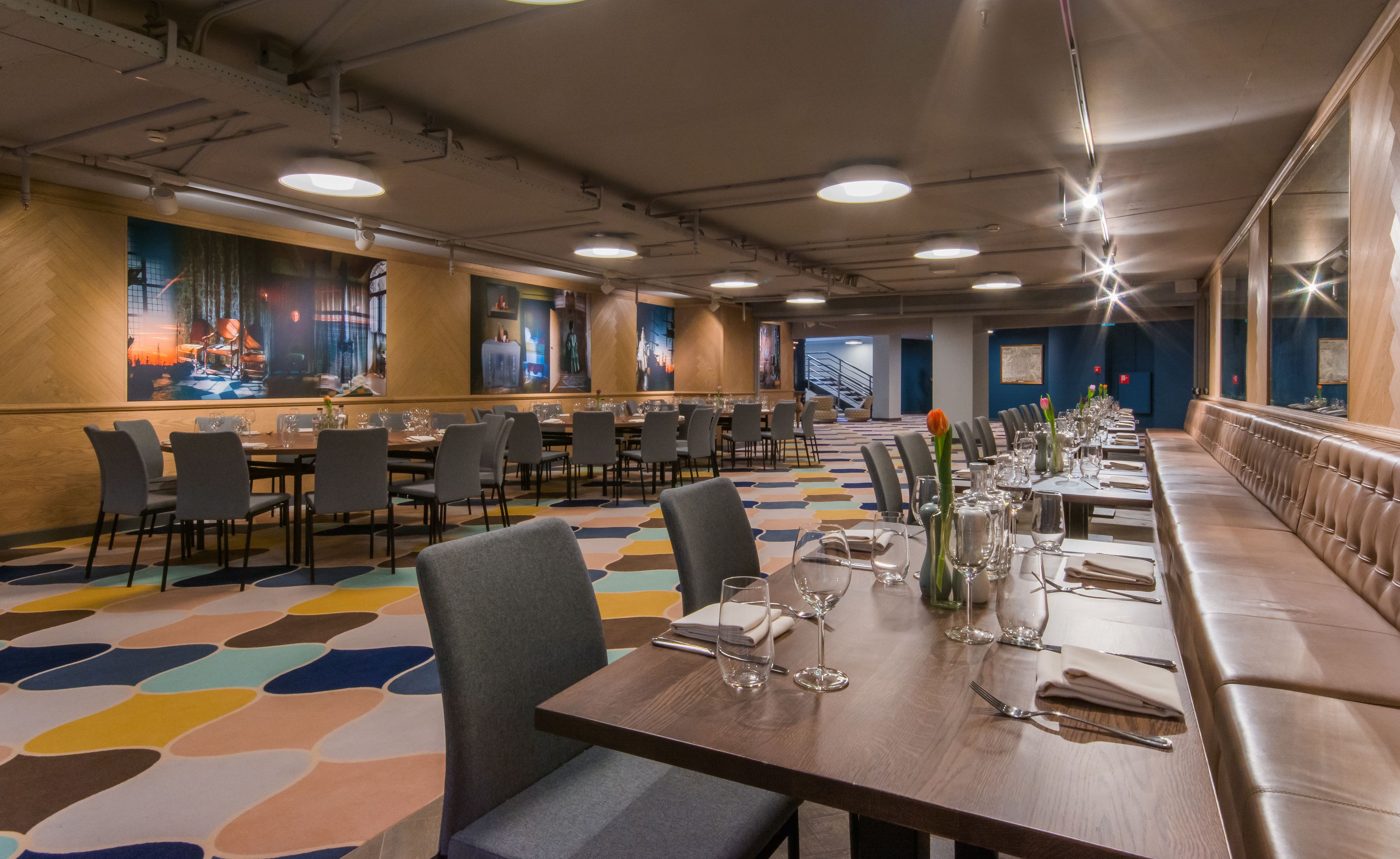 A long dining area with multiple tables and chairs, a colorful carpet, and artwork on the walls.