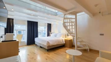 A modern hotel room with a large bed, a desk, a chair, a small table, and a staircase.