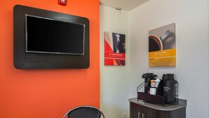 A hotel room with a flat-screen TV mounted on the wall, a coffee station with a coffee maker and cups, and a poster with the text "AT YOUR SERVICE". - Motel 6 Tucker, GA - Atlanta Northeast (Tucker)