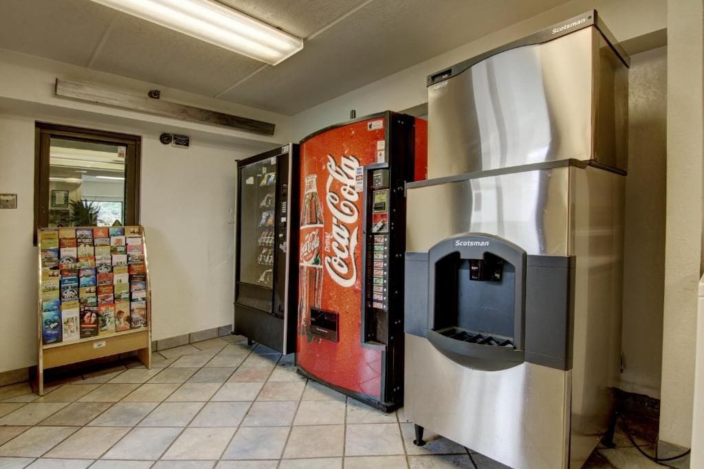 A vending machine with a Coca-Cola logo, a refrigerator, and a book rack in a hallway.