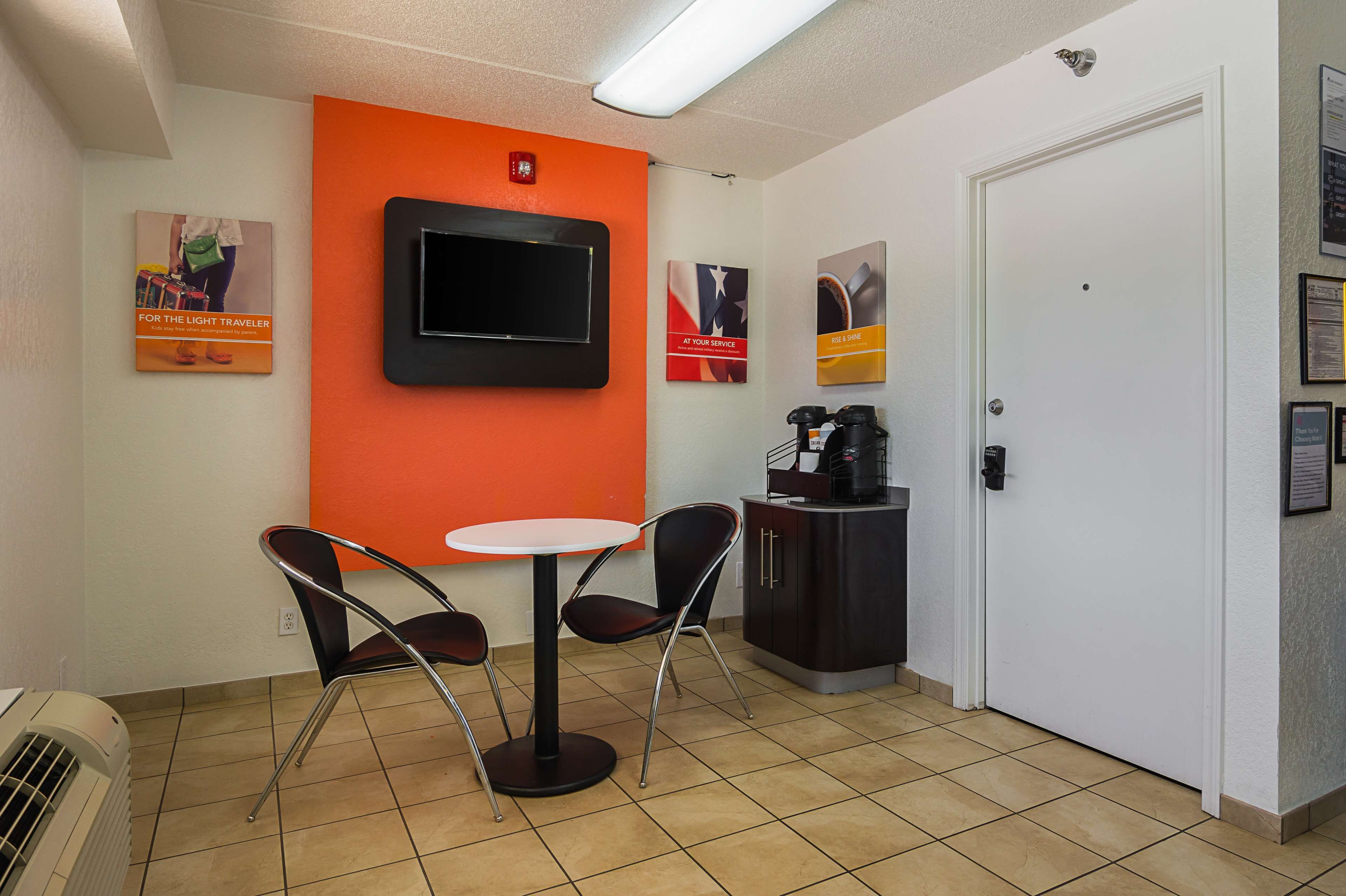 A small, brightly colored room with a white door, a black TV mounted on an orange wall, a small table with two chairs, and a coffee machine.