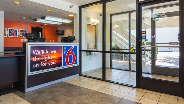 A motel lobby with a reception desk, a large sign reading "We'll leave the light on for you.", and a glass door leading outside. - Motel 6 Tucker, GA - Atlanta Northeast (Tucker)