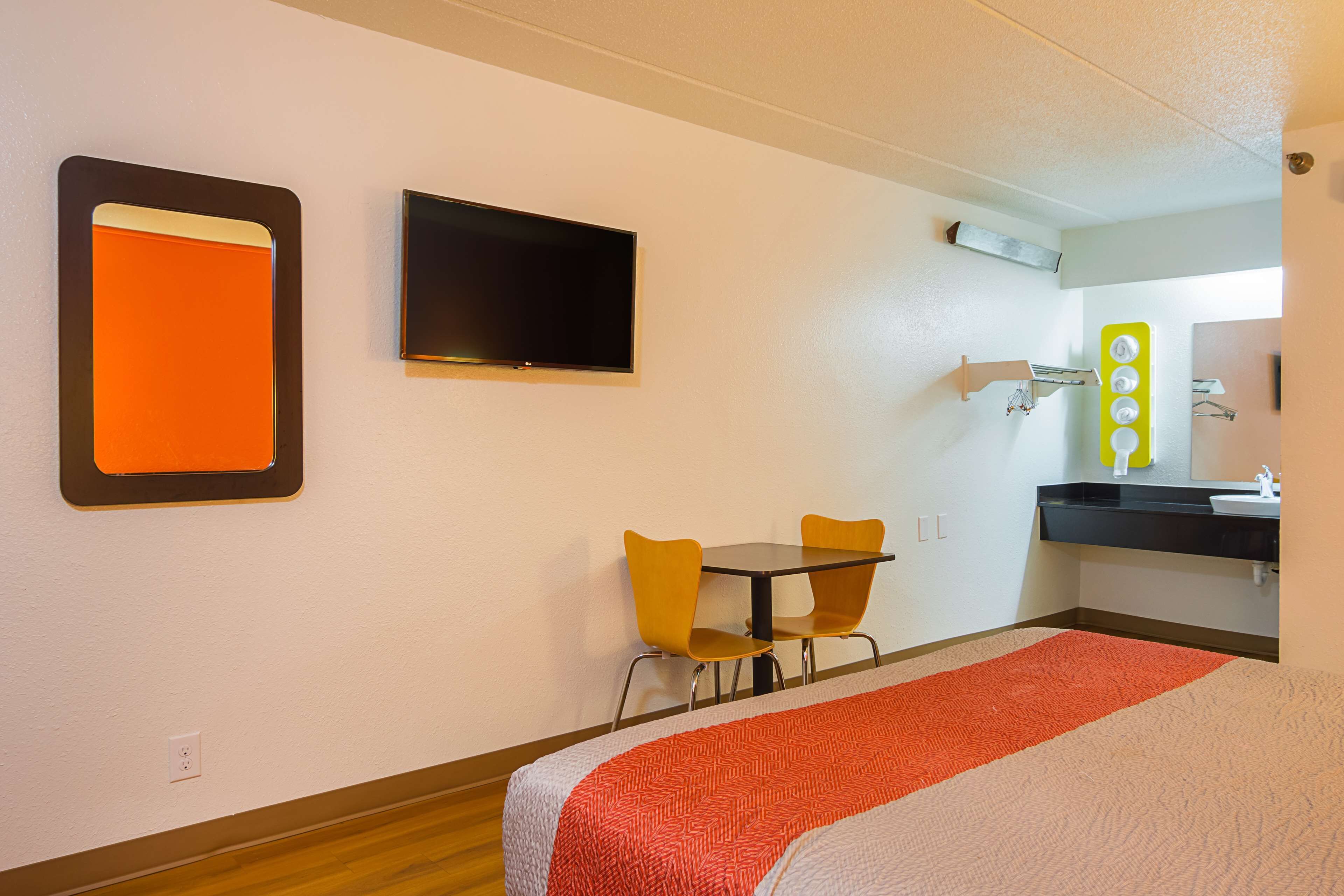 Habitación de hotel con cama, televisor, escritorio y dos sillas, y baño con espejo y lavamanos.