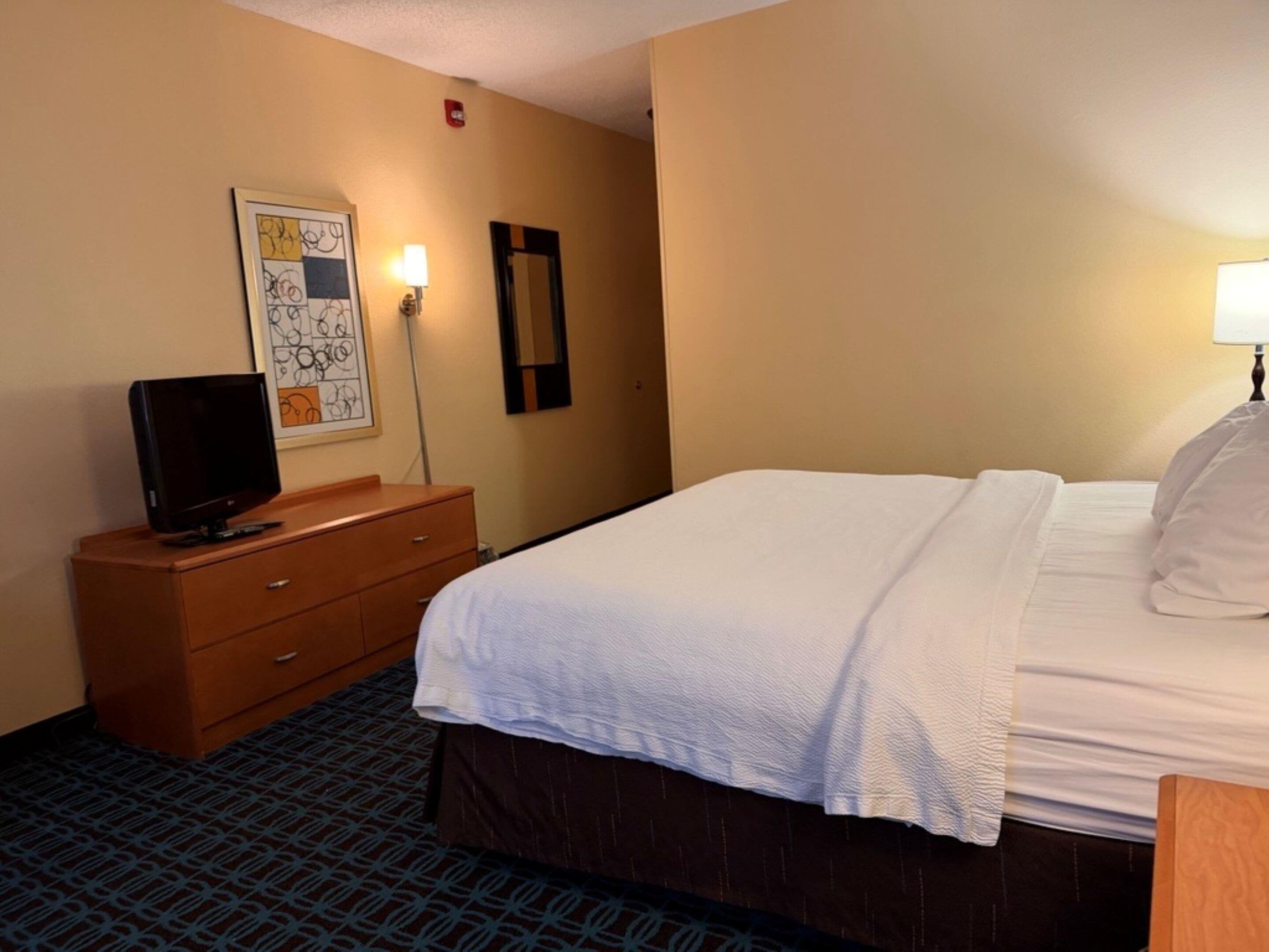 Une chambre d’hôtel avec un lit, un téléviseur posé sur une commode en bois, une lampe fixée au mur et une œuvre d’art abstraite encadrée.