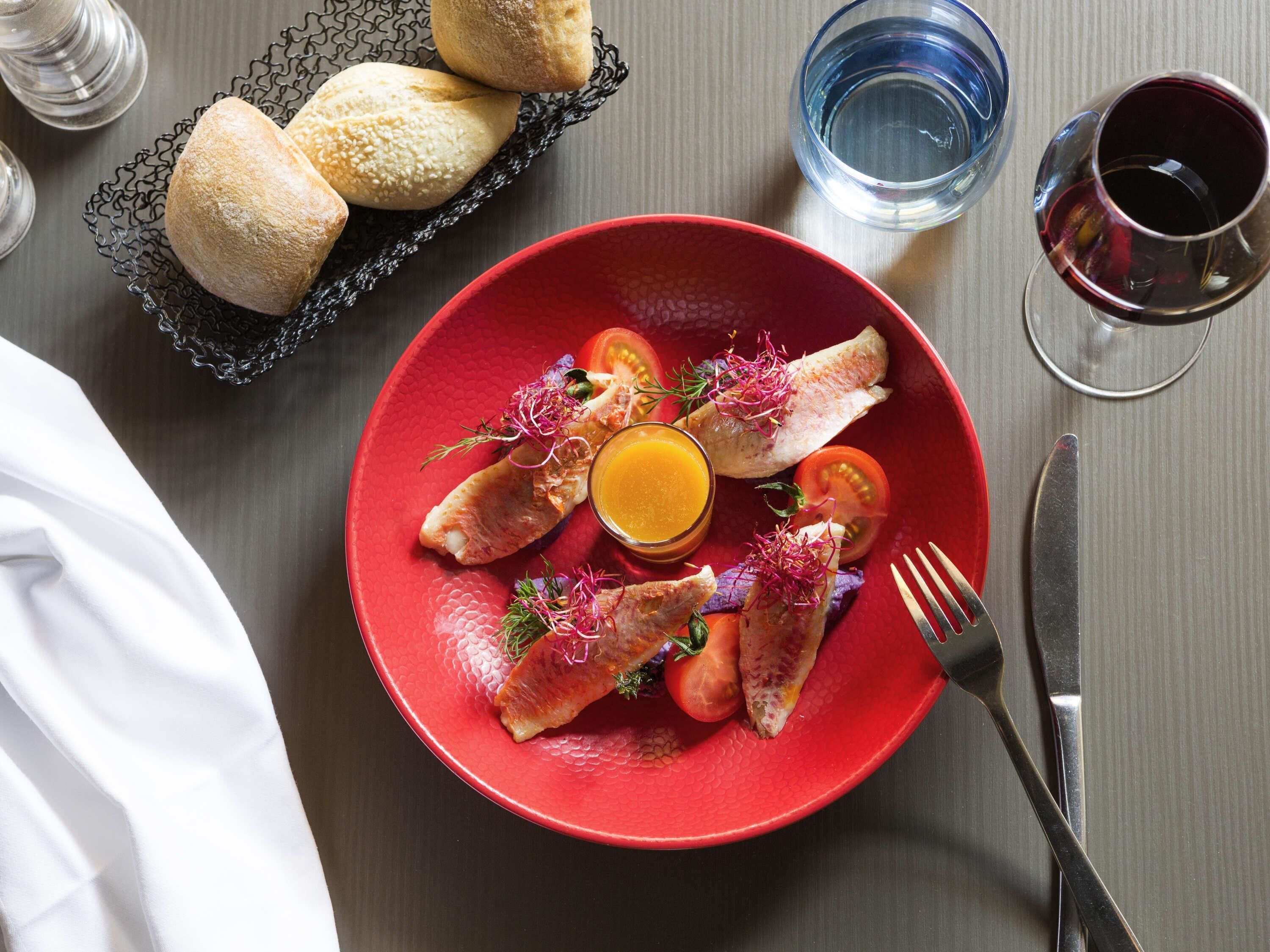 Une assiette rouge contenant du poisson, des tomates et un verre de jus d’orange, posée sur une table.