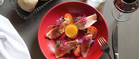 Une assiette rouge contenant du poisson, des tomates et un verre de jus d’orange, posée sur une table.