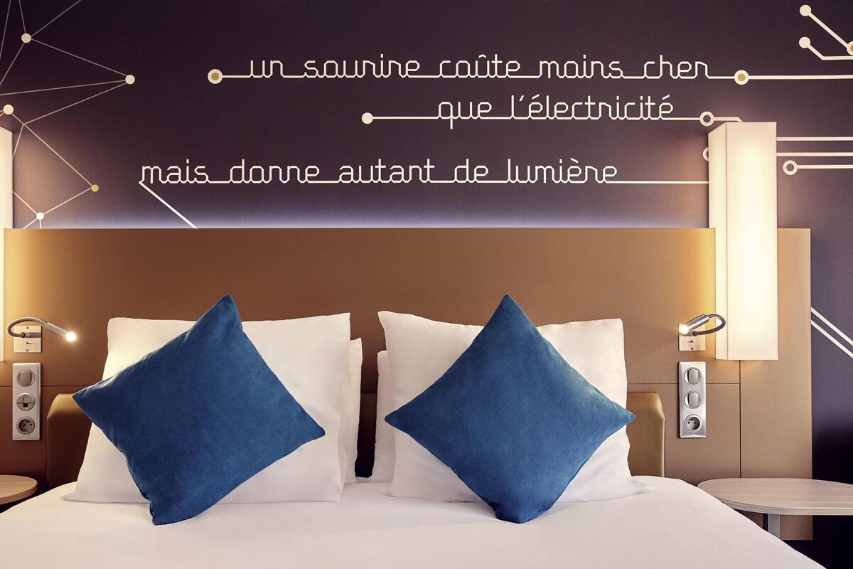 Quarto de hotel moderno com uma cama bem arrumada, duas almofadas azuis, luminárias de cabeceira e um aplique de parede.