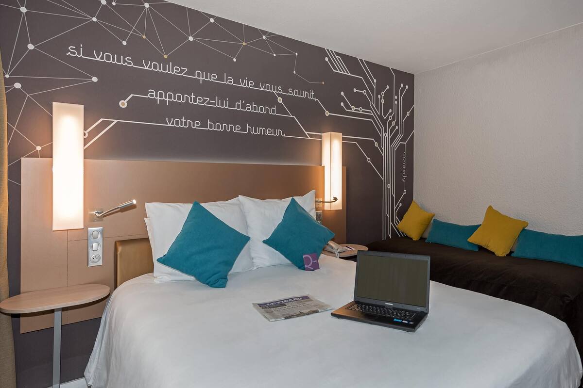 Quarto de hotel com uma cama, um laptop, uma mesa de cabeceira e uma parede com um design geométrico.