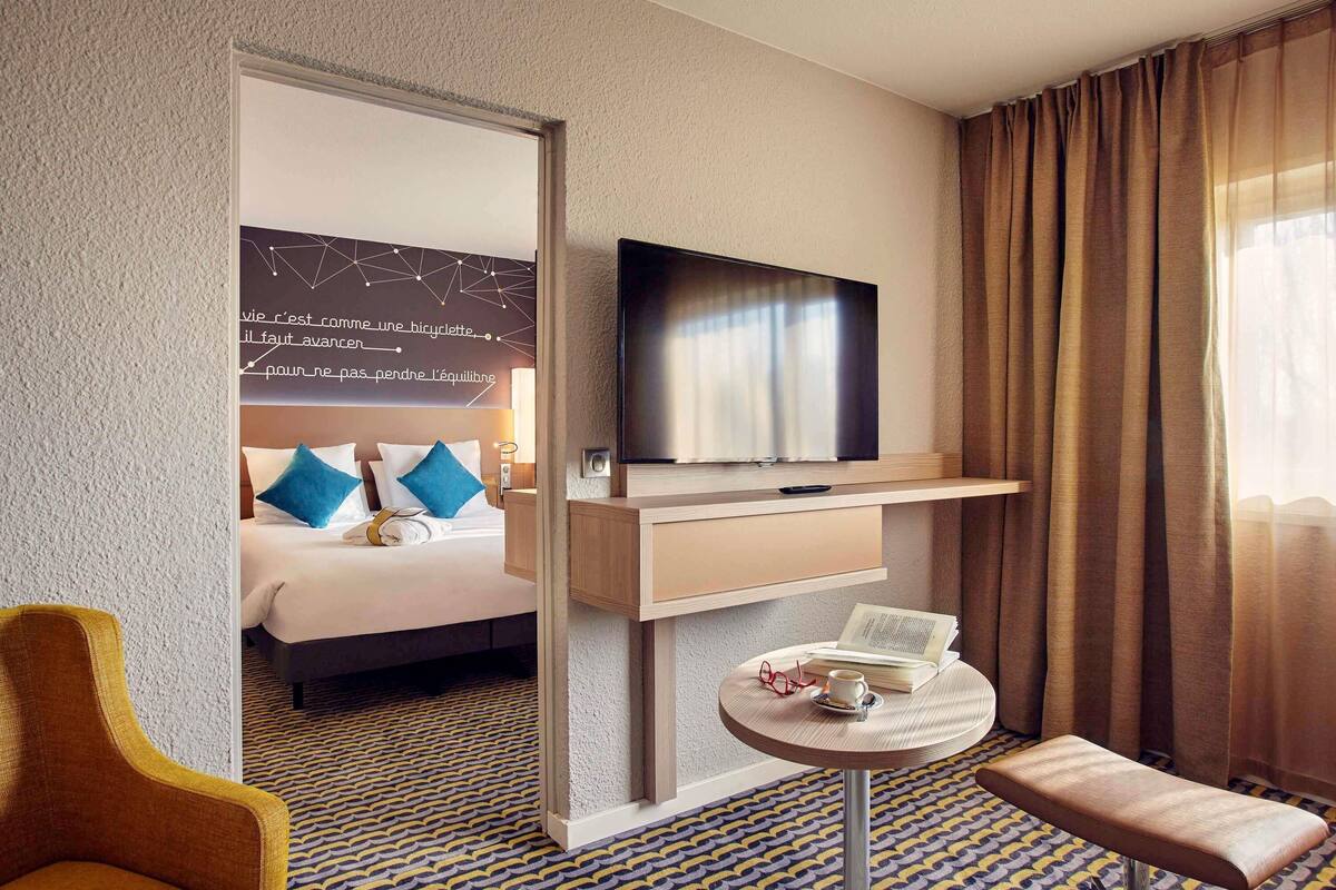 Quarto de hotel com cama, televisão, pequena mesa com um livro e copos, uma cadeira e tapete estampado.