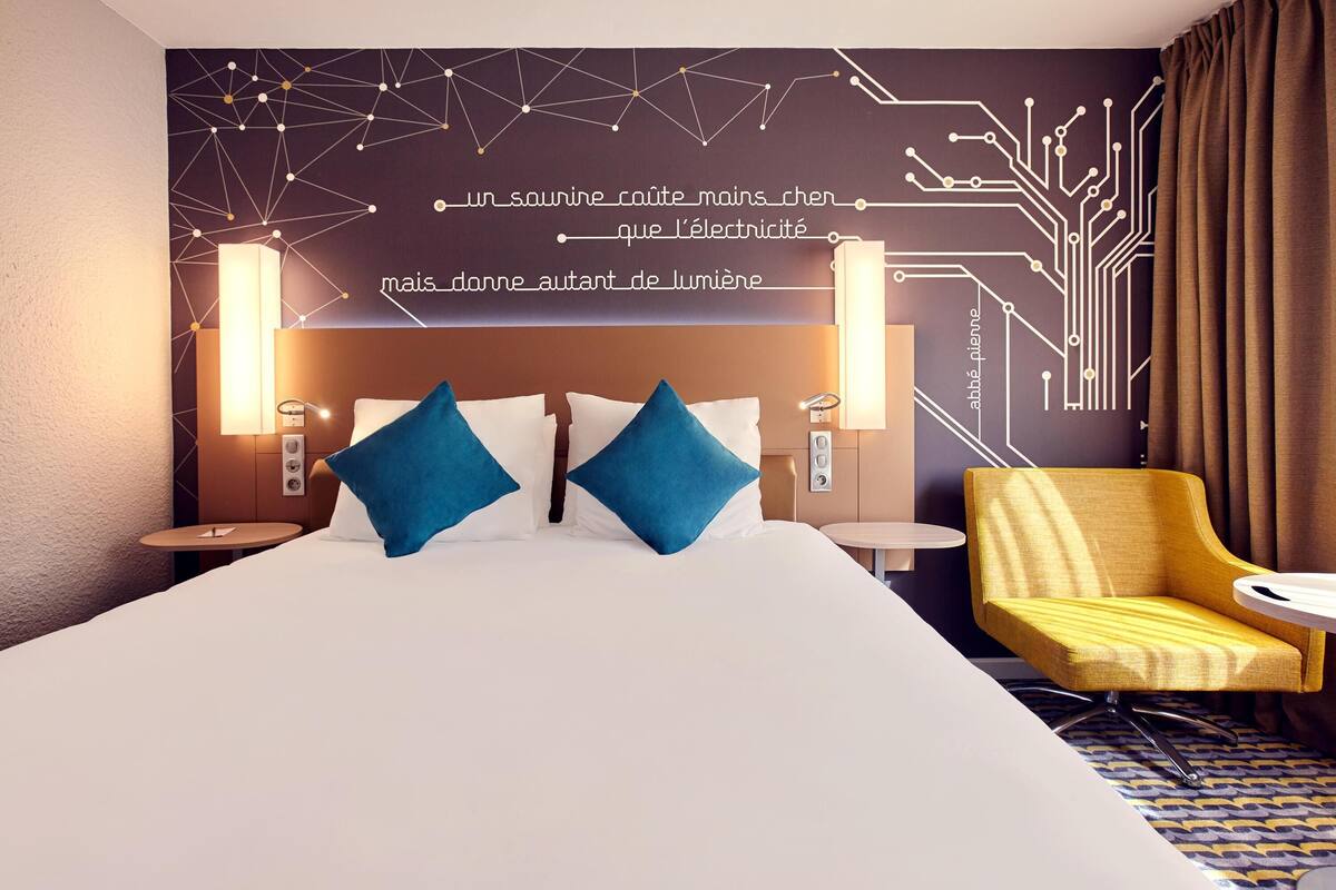 Quarto de hotel com uma cama grande, duas almofadas azuis, uma poltrona amarela e uma parede com padrões geométricos.