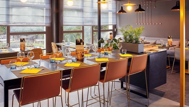 A modern dining area with a long table, orange chairs, and pendant lighting. - Mercure Paris Sud Les Ulis (Les Ulis)