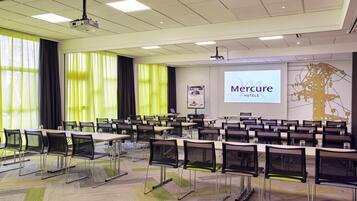 Un salón de conferencias con filas de mesas y sillas, un proyector y una pantalla mostrando 'Mercure Hotels'.