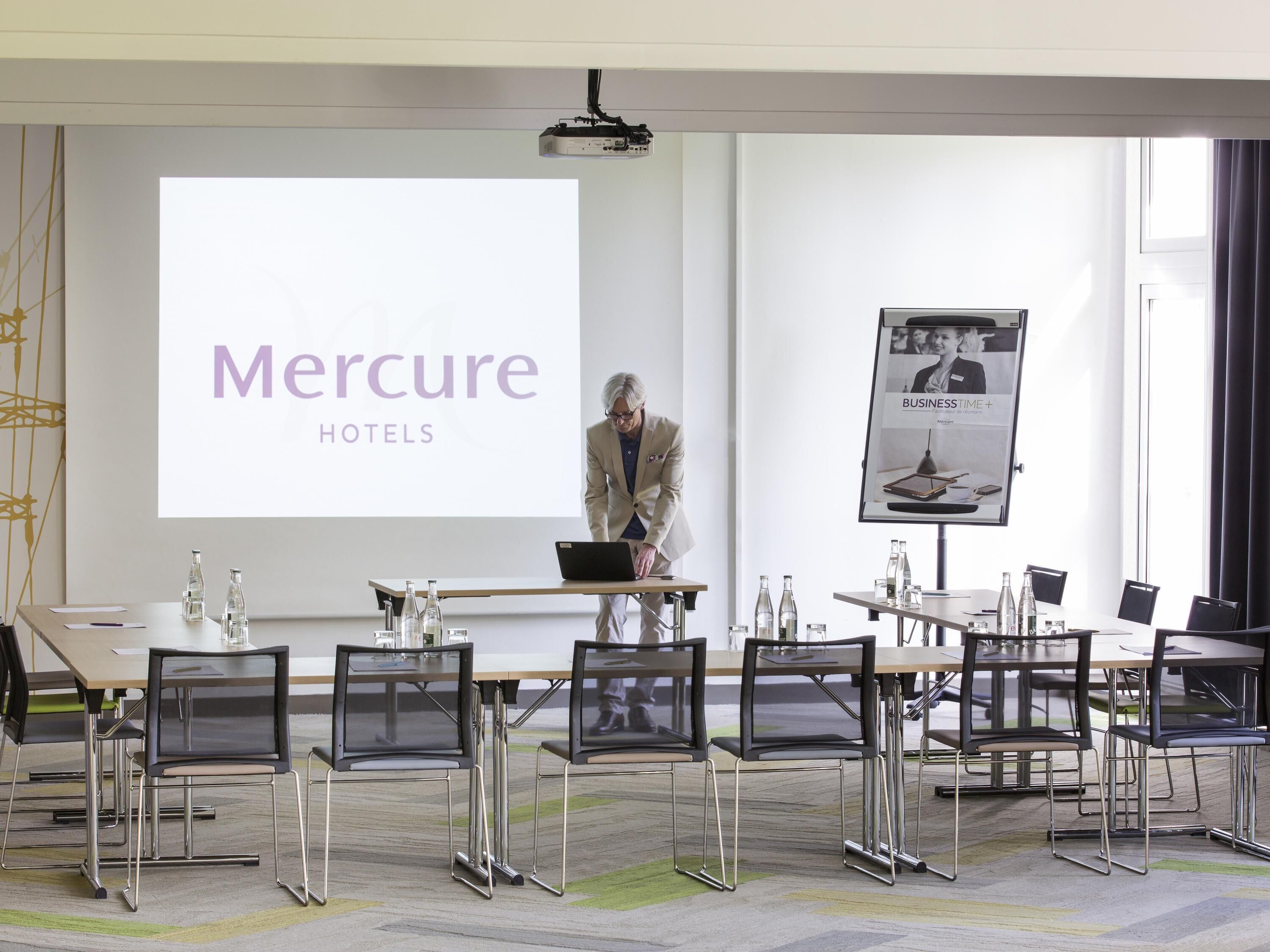 A conference room with a presentation screen displaying 'Mercure HOTELS,' a projector, and a poster on the wall.