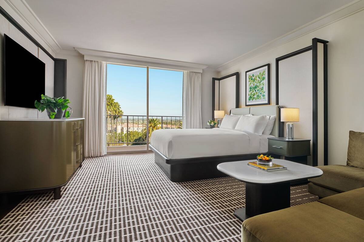 A hotel room with a large bed, a sofa, a small table, a TV, a plant, and a balcony with a view.
