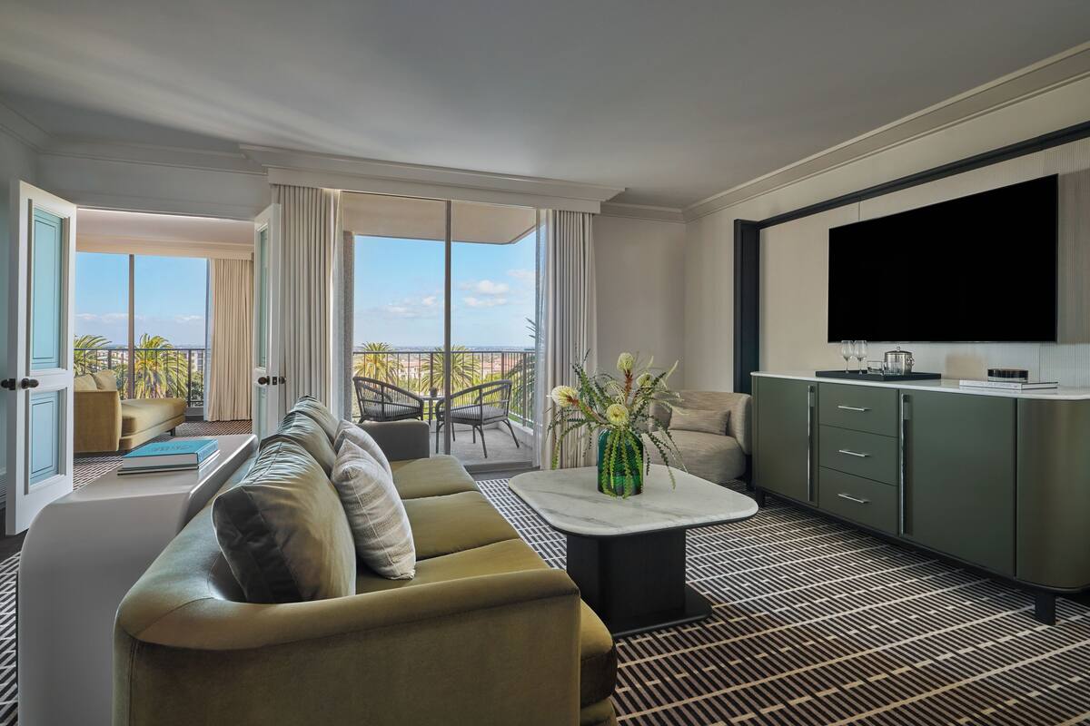 A modern hotel room with a sofa, a coffee table with a vase, a TV, and a balcony with a view.