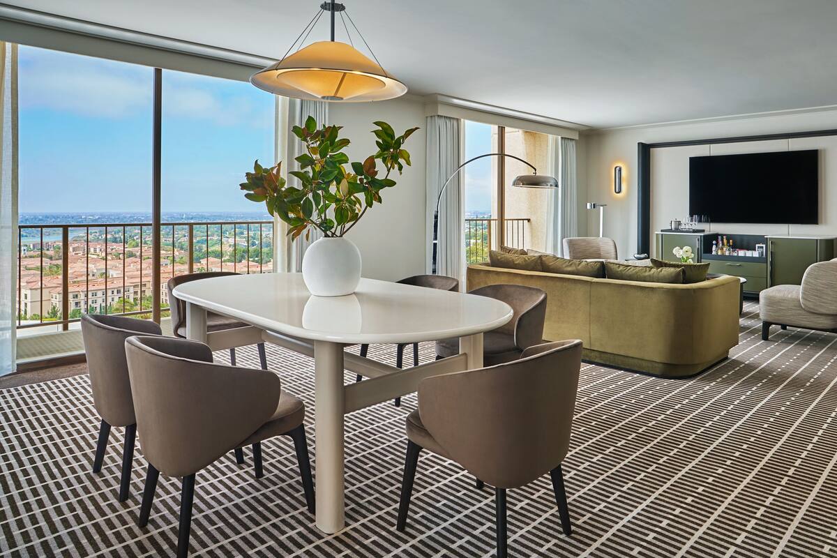 A modern living room with a dining area, large windows, a flat-screen TV, and a view of the cityscape.