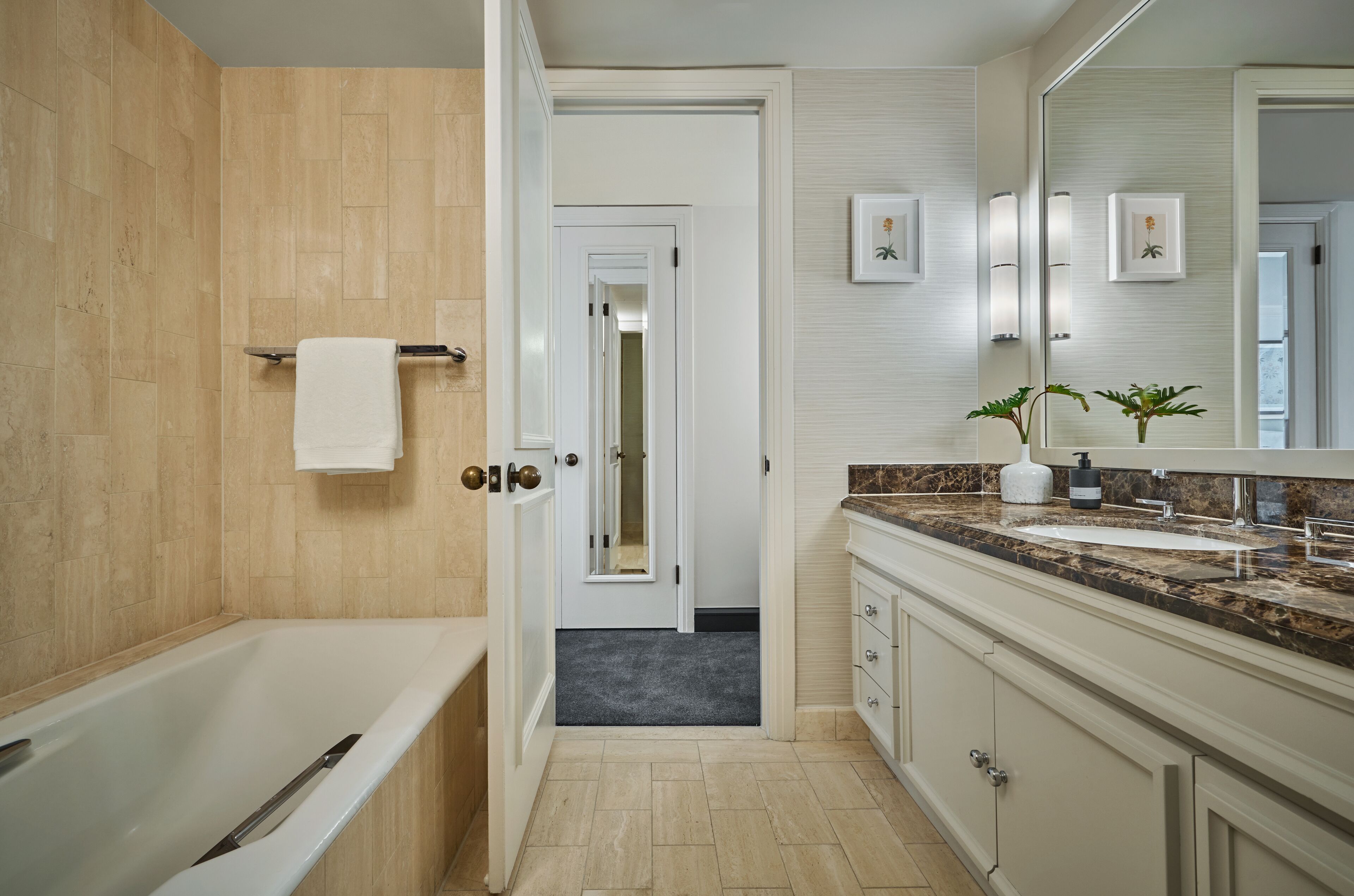 Un baño moderno con bañera, lavamanos y espejo. Se ve una puerta que conduce a otra habitación.