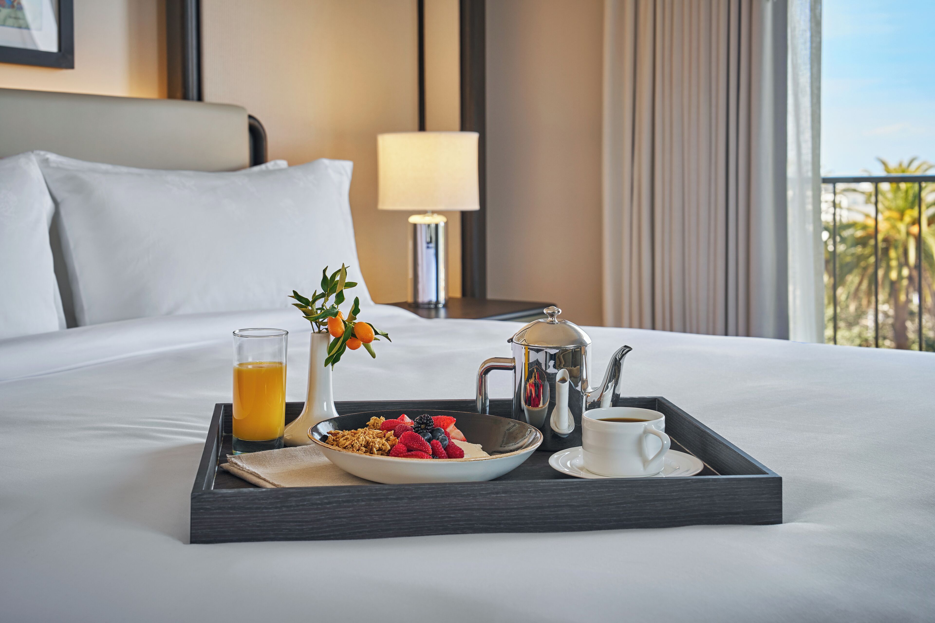 A neatly made breakfast tray on a hotel bed with a view of palm trees.