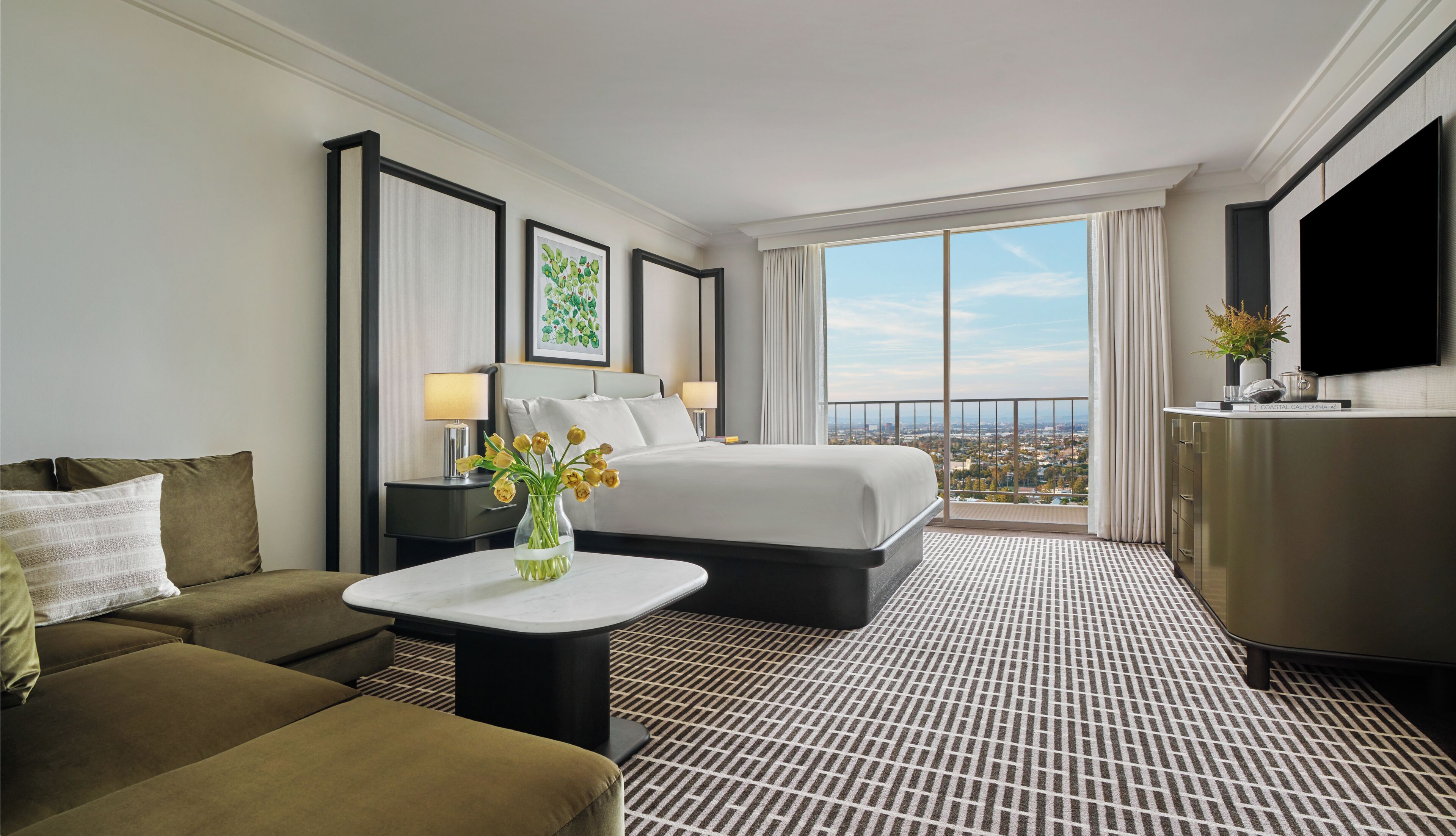 Ein Hotelzimmer mit einem Bett, einem Sofa, einem kleinen Tisch mit Blumenvase, einem Fernseher und einem großen Fenster mit Blick auf die Stadt.