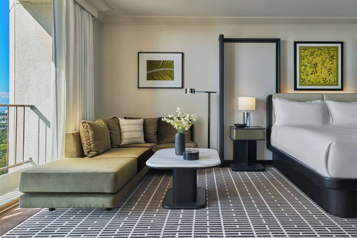 A hotel room with a sofa, a small table with a vase, a bed, and two framed pictures on the wall.