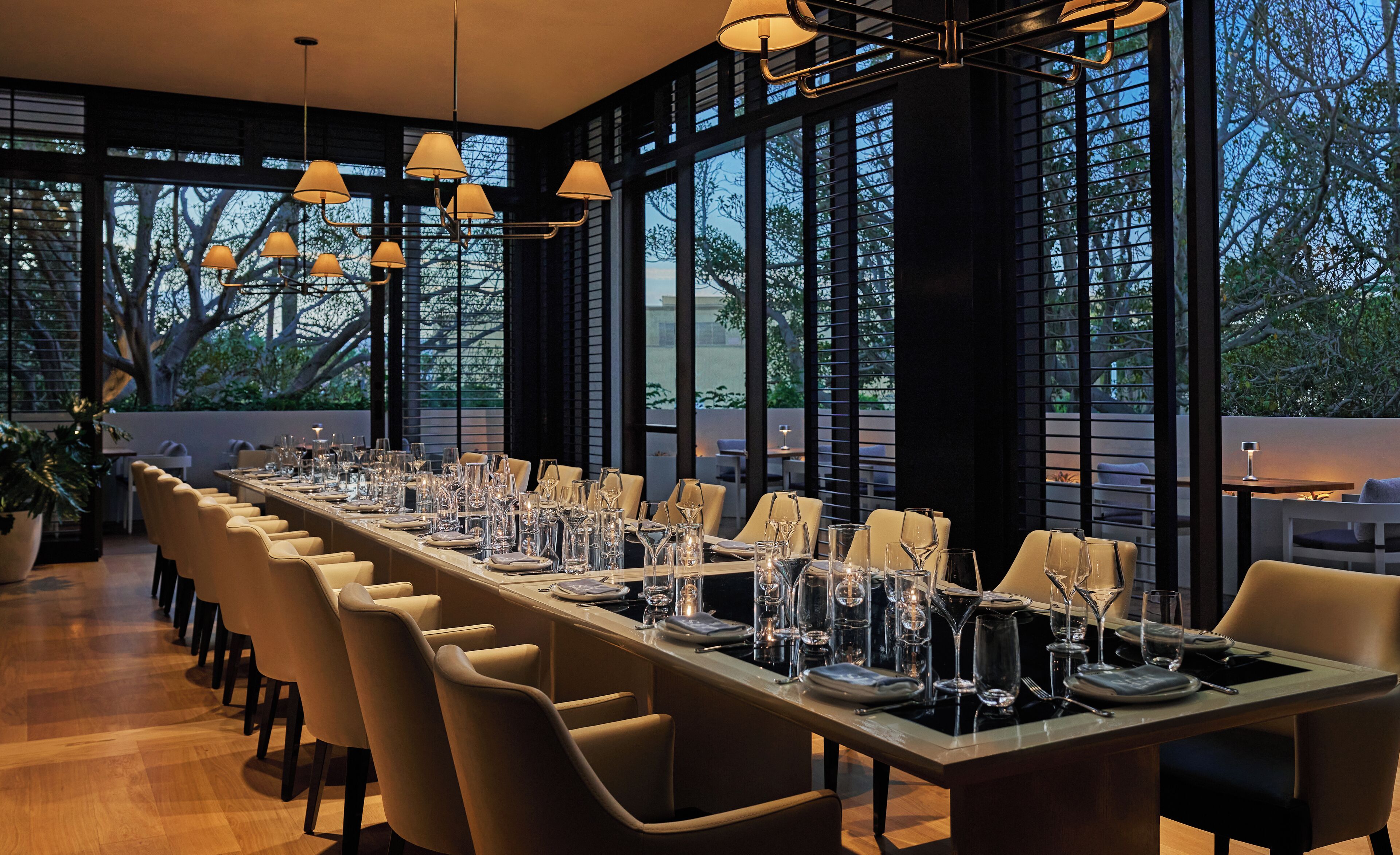A long dining table set with chairs, large windows, and hanging lamps.
