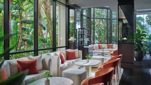 A modern indoor space with a sofa, ottomans, and chairs arranged around a central table. Large windows provide views of lush greenery outside. - Pendry Newport Beach (Newport Beach)