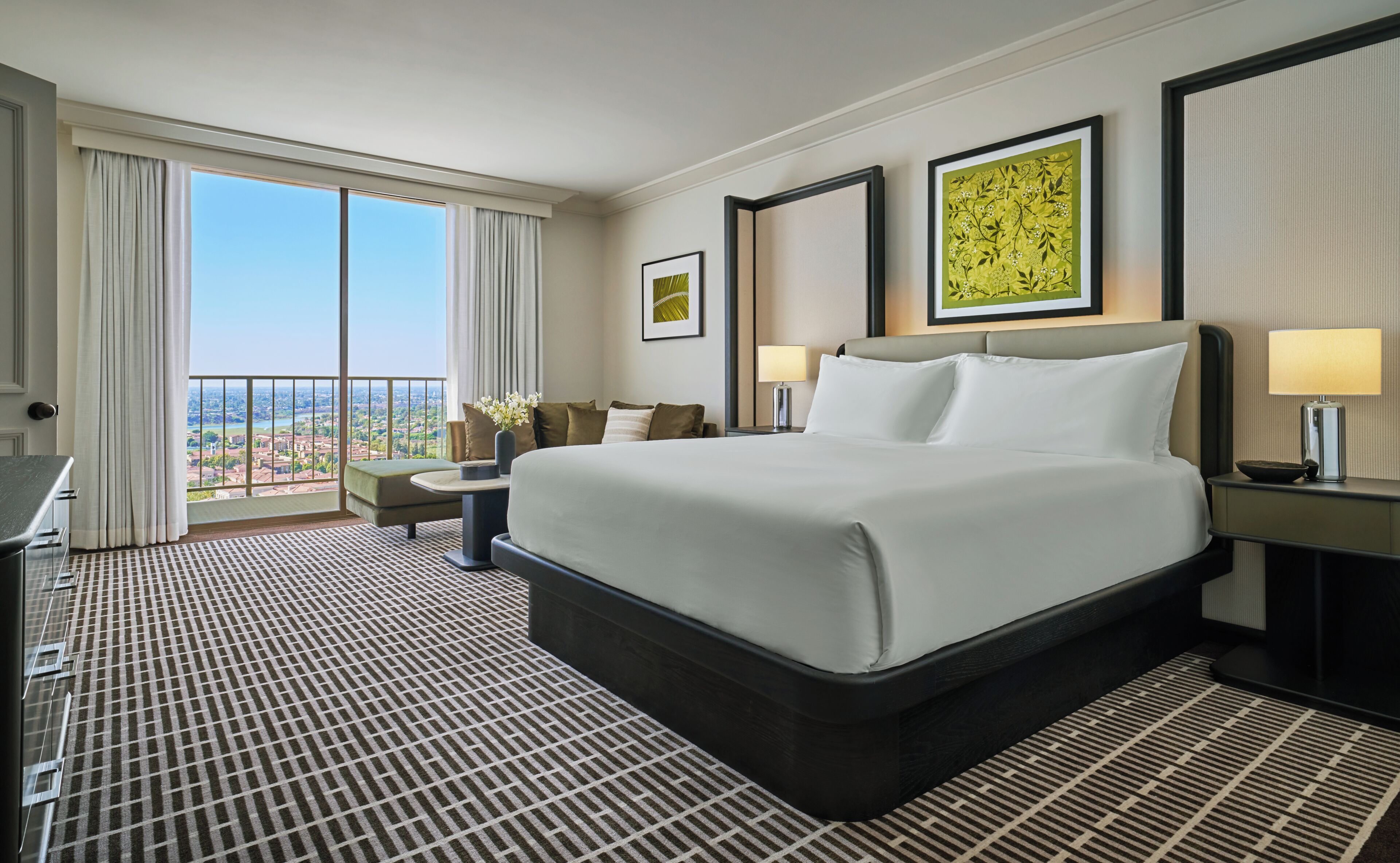 A hotel room with a large bed, a desk, a chair, a view of the city, and artwork on the walls.