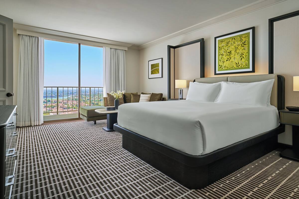 A hotel room with a large bed, a desk, a chair, a view of the city, and artwork on the walls.
