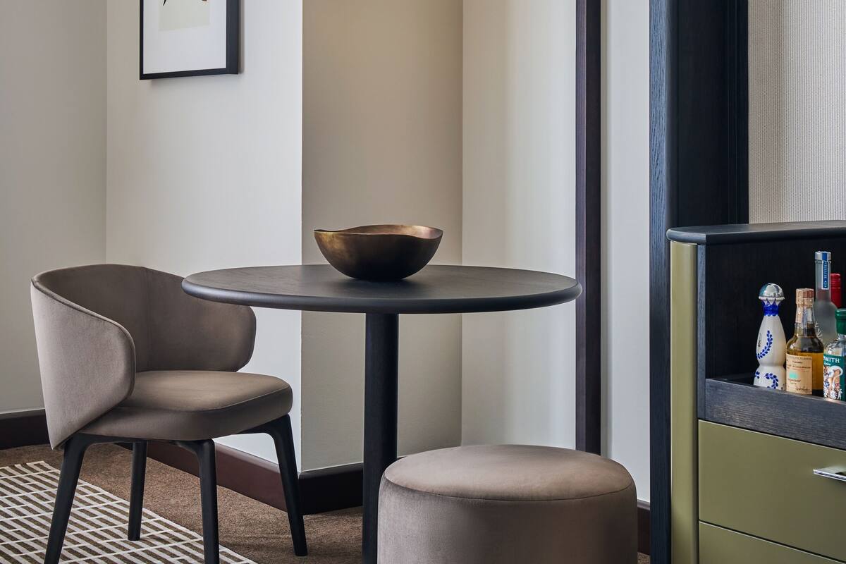 A modern hotel room with a round table, a chair, a small stool, a hanging lamp, a framed artwork, and a minibar.