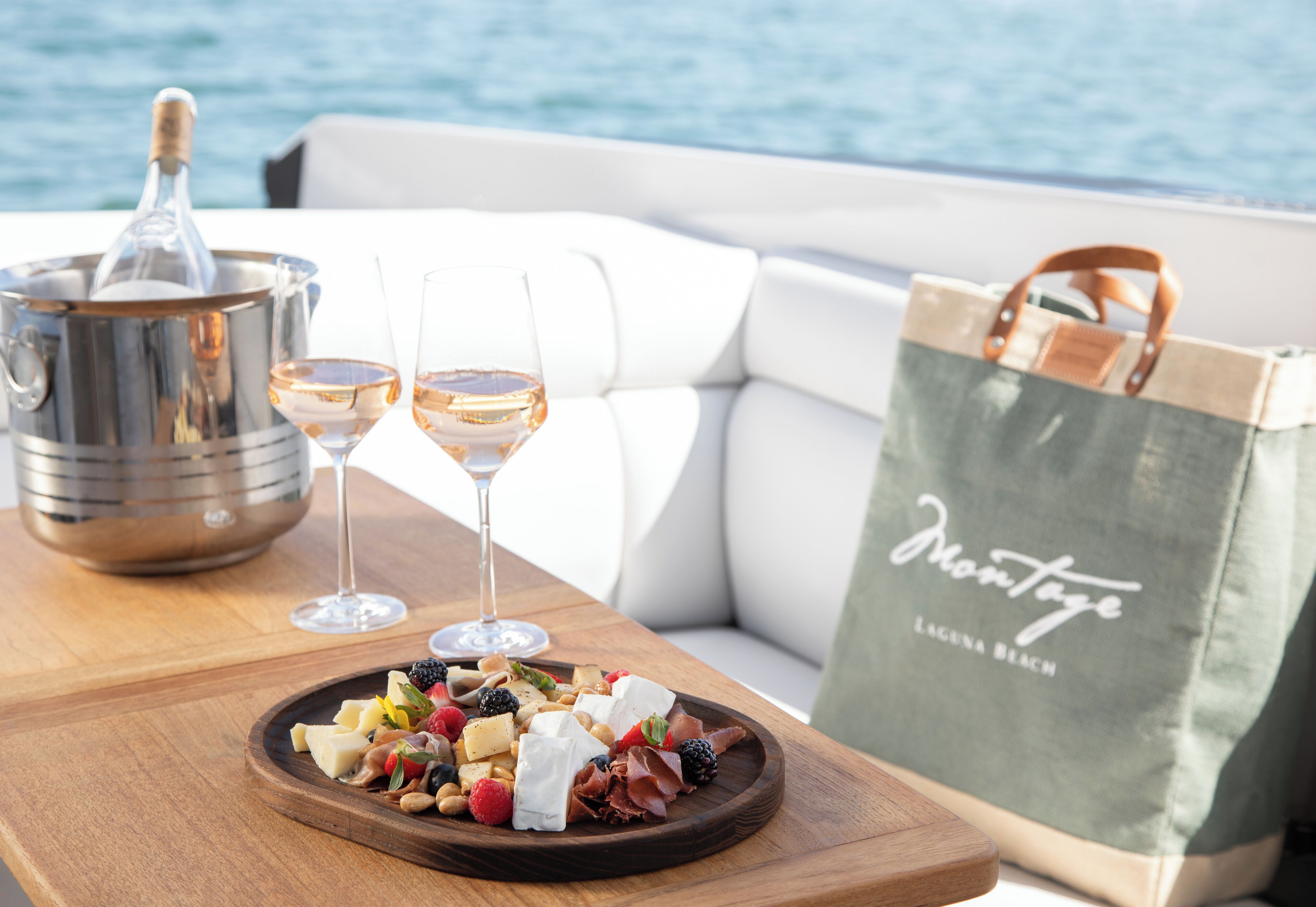 A wooden table with a cheese platter, two glasses of wine, and a bottle of champagne on a boat deck.