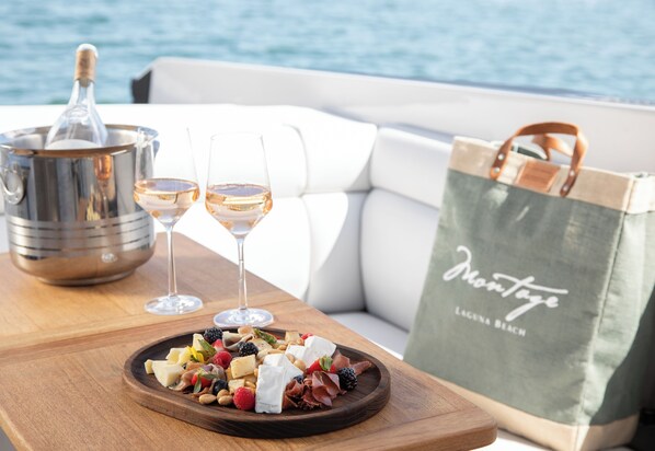 A wooden table with a cheese platter, two glasses of wine, and a bottle of champagne on a boat deck.