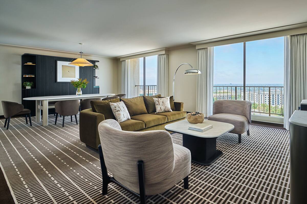 A modern hotel room with a sofa, armchairs, a dining table, and a balcony with a city view.
