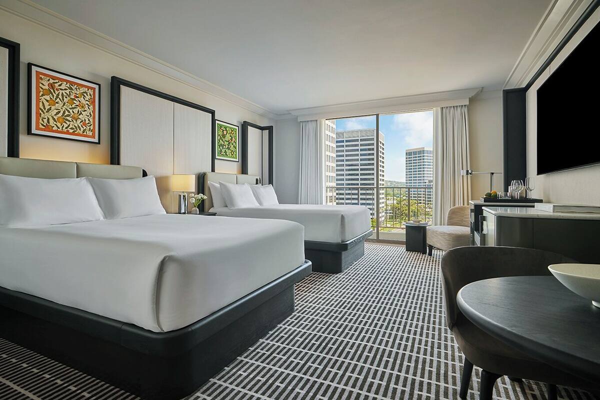 A hotel room with two beds, a dining area, a TV, and a balcony with a city view.