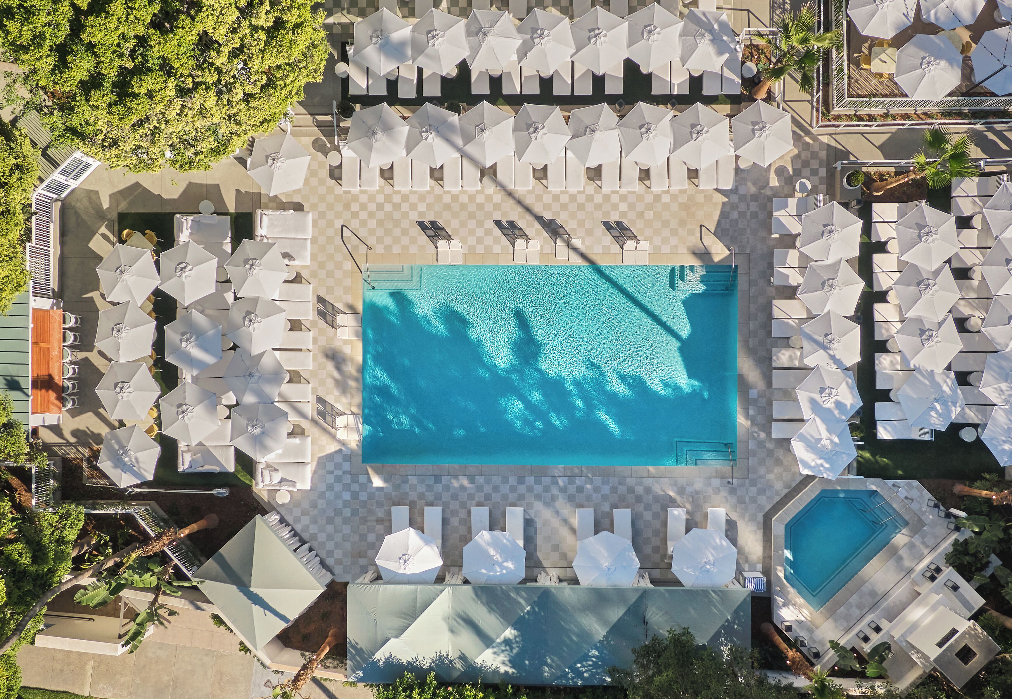 An aerial view of a pool area with multiple sun loungers and umbrellas, surrounded by trees and a checkered floor pattern.