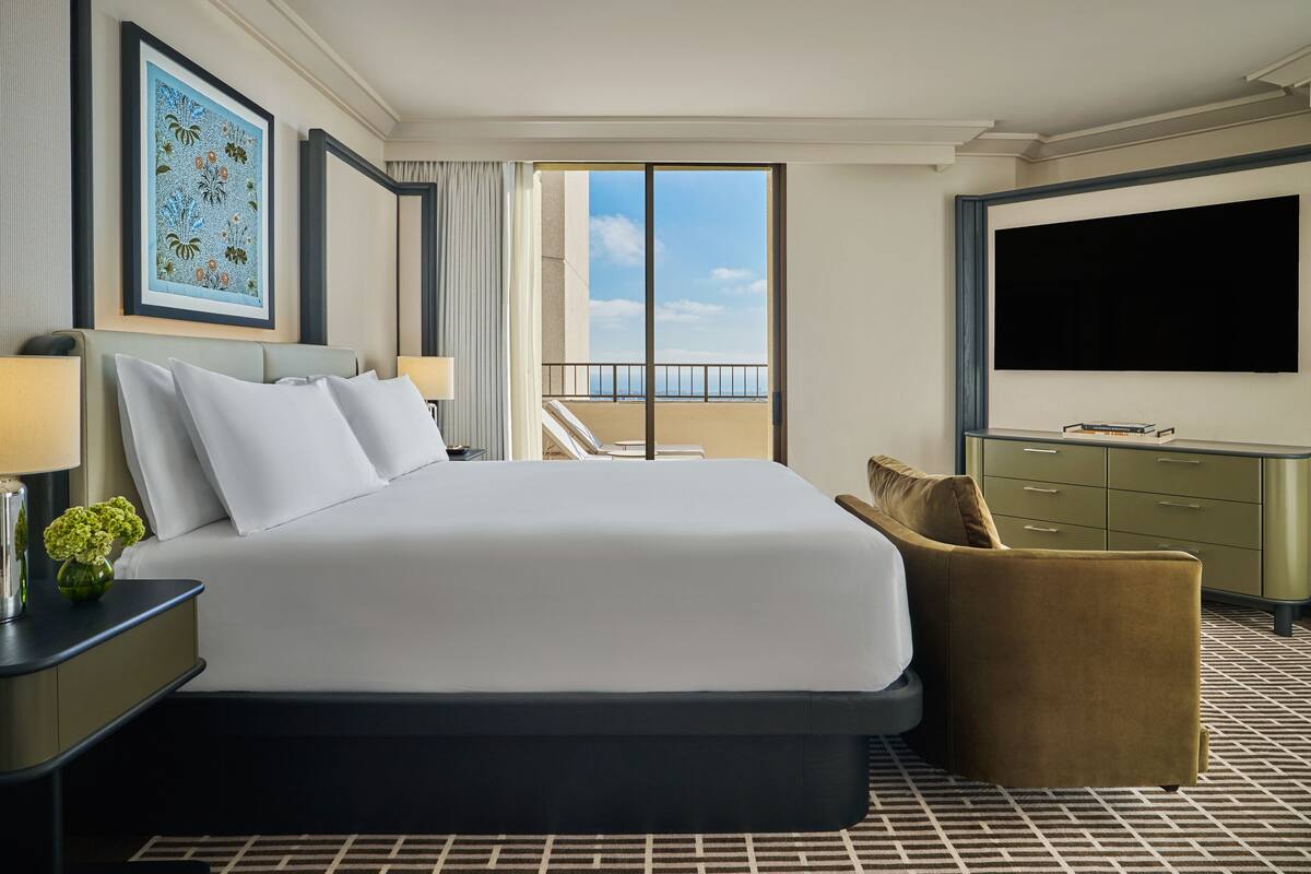 A hotel room with a large bed, a TV, a desk with a lamp, a chair, and a view of the sky through the balcony doors.