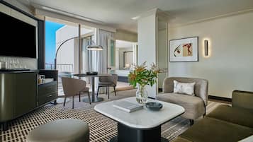 A modern hotel room with a dining area, a living room with a sofa and armchair, a flat-screen TV, and a large window with a view.