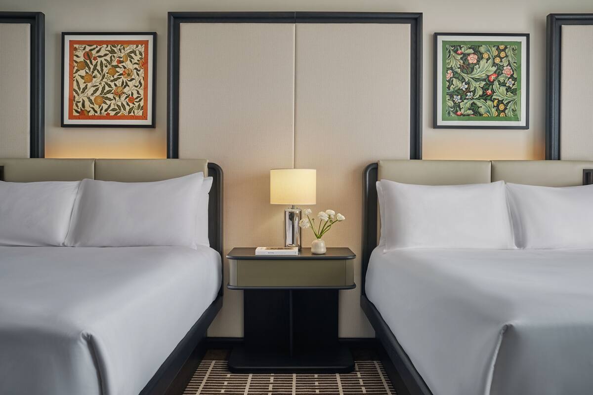 A hotel room with two beds, a nightstand with a lamp, and two framed floral artworks on the wall.