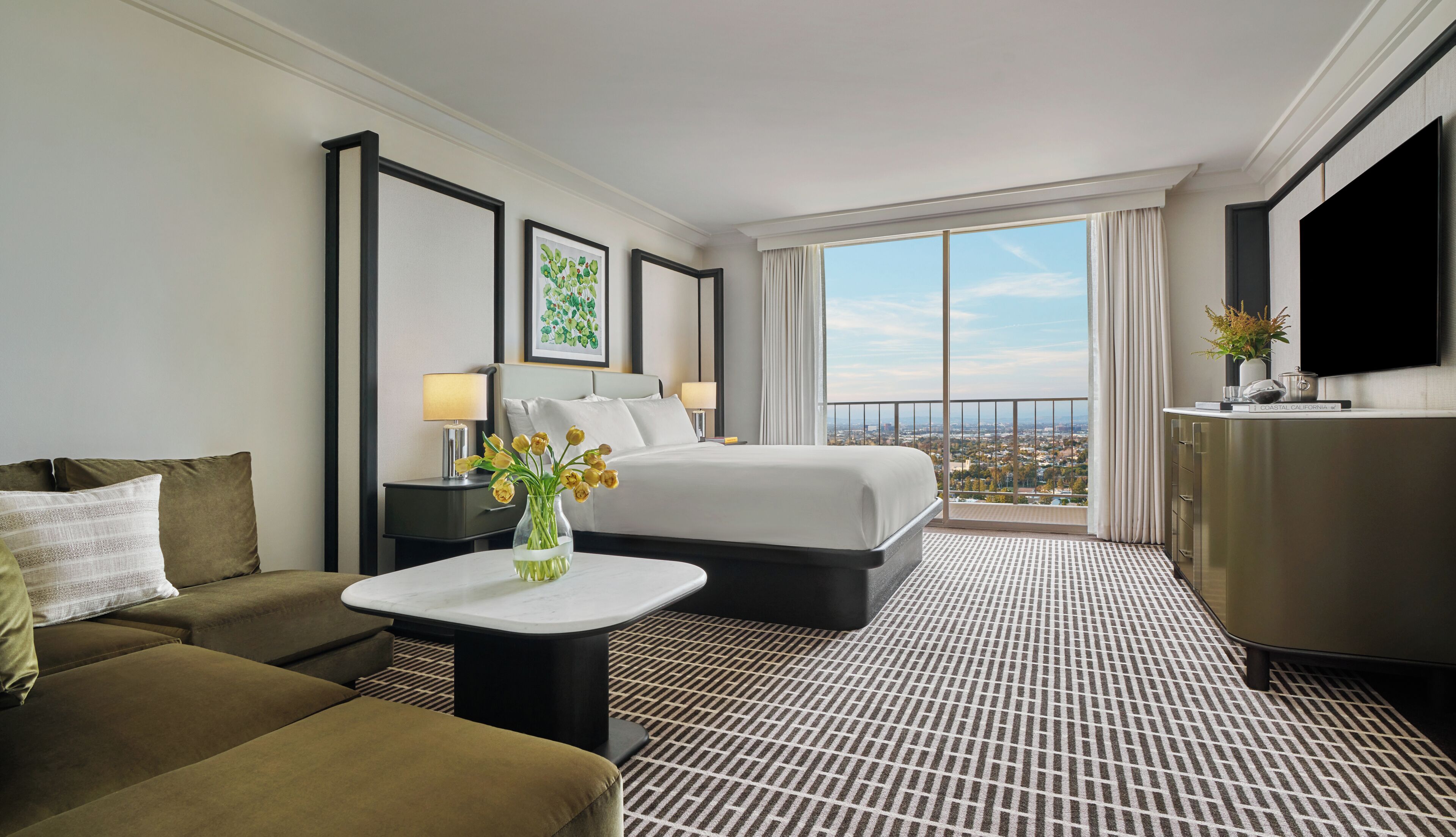 A hotel room with a bed, a sofa, a small table with a vase of flowers, a TV, and a large window with a city view.