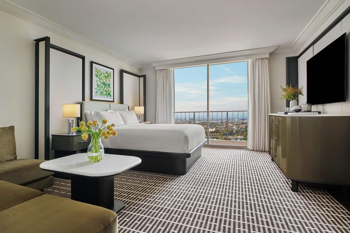 A hotel room with a bed, a sofa, a small table with a vase of flowers, a TV, and a large window with a city view.
