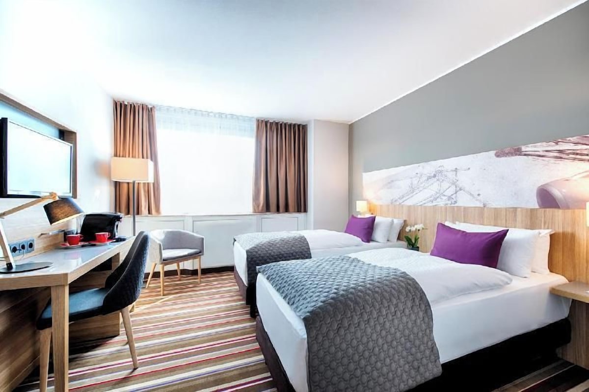Habitación de hotel con dos camas, un escritorio con computadora, una silla, un televisor y una ventana con cortinas.