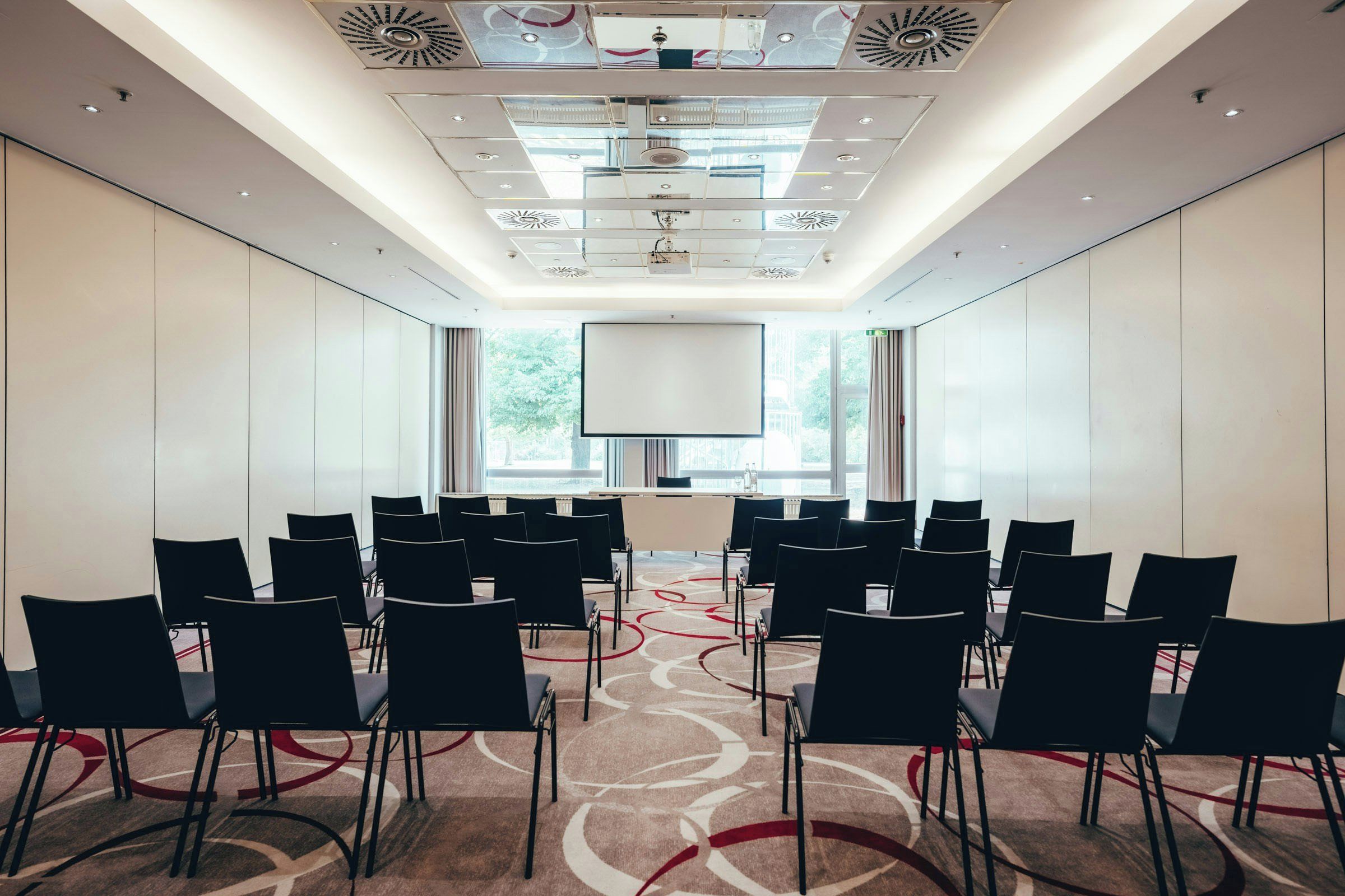 Ein Konferenzraum mit Stuhlreihen, einer großen Leinwand und einem gemusterten Teppich.