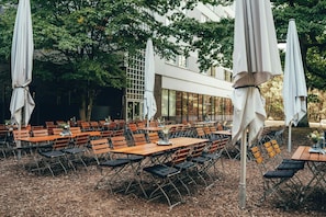 Un área de comedor al aire libre con mesas y sillas, rodeada de árboles y un edificio con ventanas amplias.