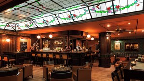 Zona de bar con techo alto, vidrieras de colores y un cantinero. Varias áreas de estar con mesas y sillas.