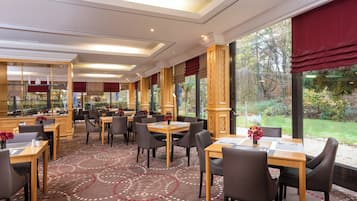 Une salle à manger d’hôtel avec des tables et des chaises, de grandes fenêtres et un tapis à motifs.