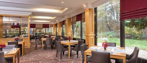 Een restaurantgedeelte in een hotel met tafels en stoelen, grote ramen en een kamerbreed tapijt.
