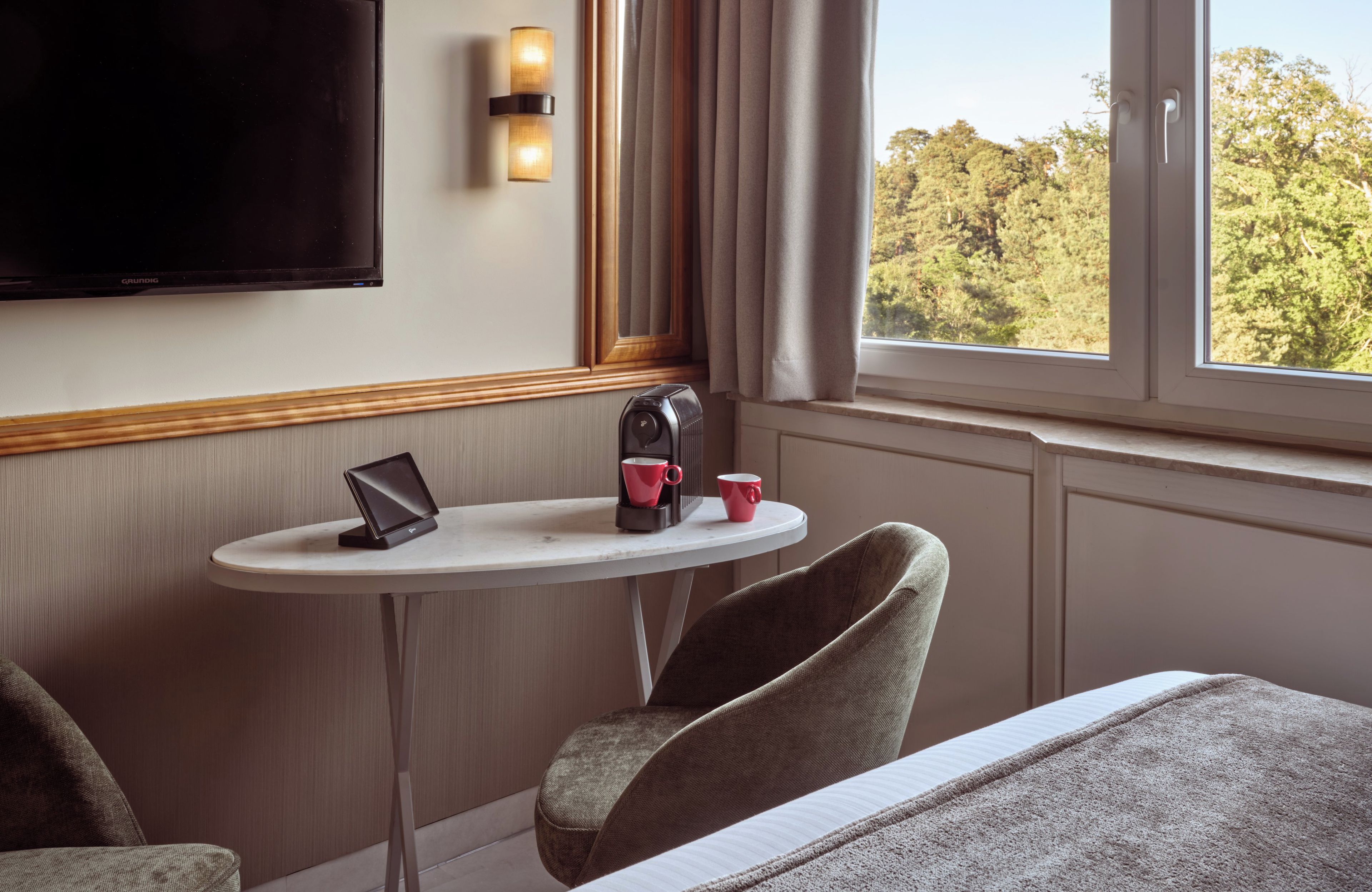 Een hotelkamer met een bed, een klein tafeltje met een laptop, een koffiezetapparaat en een kopje. Er is een tv aan de muur bevestigd en een raam met gordijnen.