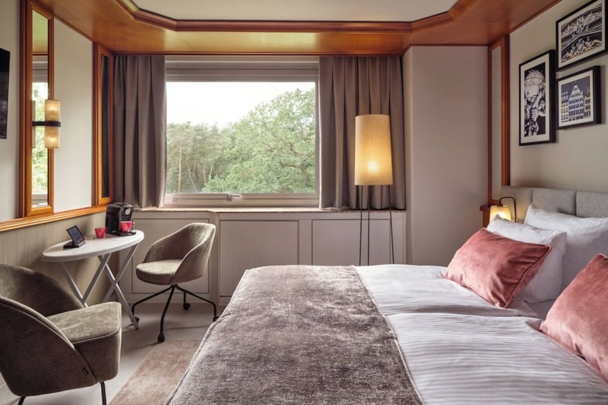 Habitación de hotel con cama, escritorio, dos sillas, ventana con cortinas e imágenes enmarcadas en la pared.
