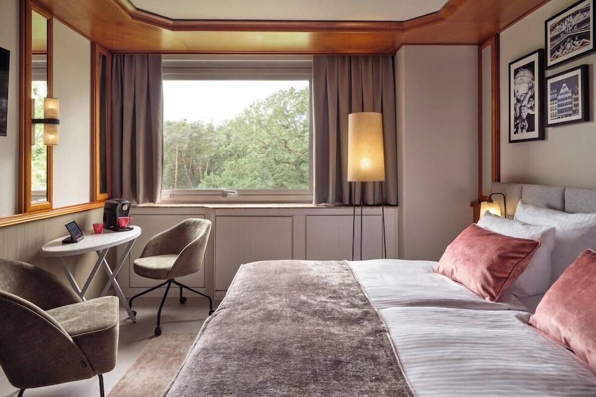 Habitación de hotel con cama, escritorio, dos sillas, ventana con cortinas e imágenes enmarcadas en la pared.
