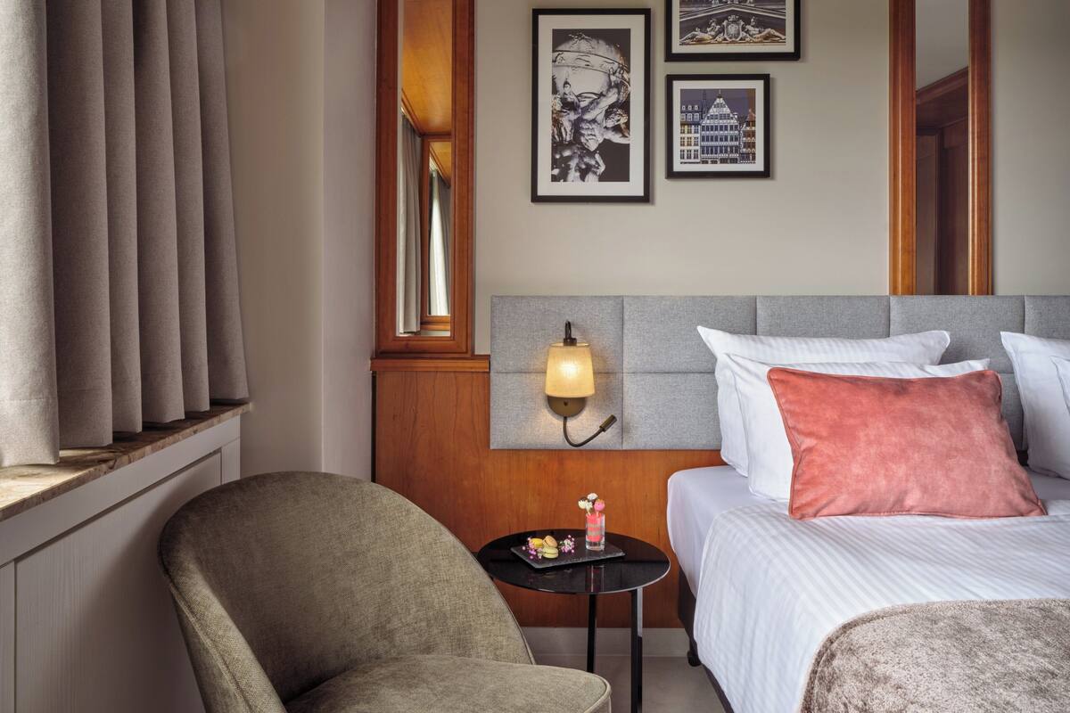 Habitación de hotel con cama, mesita de noche, una silla, una pequeña mesa con lámpara y cuadros en la pared.